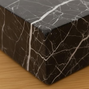 May include: Close-up of a black marble block with white veining. The polished surface reflects light, highlighting the natural patterns. The corner of the block is visible, showcasing the material's texture and color variations.