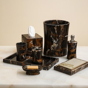 Può includere: Un set di accessori per il bagno con un motivo marmorizzato marrone e nero. Il set include un porta fazzoletti, un bicchiere, un portasapone, un vassoio, un cestino e un dosatore di sapone.