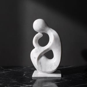Könnte beinhalten: Eine weiße Marmorskulptur einer abstrakten menschlichen Form. Die Skulptur hat eine glatte, polierte Oberfläche und ein geschwungenes Design mit einer zentralen Öffnung. Sie steht auf einem quadratischen Sockel vor dunklem Hintergrund.