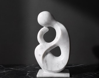Puede incluir: Escultura de mármol blanco de una forma humana abstracta. La escultura tiene un acabado liso y pulido y presenta un diseño curvo con una abertura central. Se apoya sobre una base cuadrada sobre un fondo oscuro.
