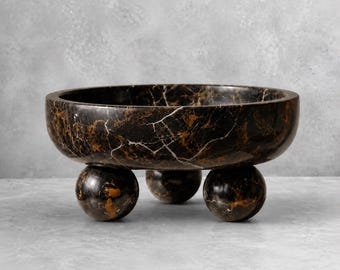 May include: A round bowl crafted from dark brown and black marble with prominent white veining. The bowl features a smooth, polished finish and is supported by three spherical feet made of the same matching marble material.