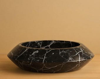 May include: A black marble bowl with white veining. The bowl has a unique, geometric shape with a wide, open top and angled sides. The bowl is made of a polished stone and sits on a wooden surface.