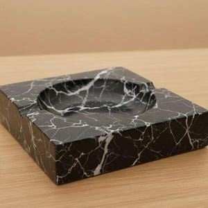 May include: A square ashtray made of black marble with white veining. The ashtray has a circular indentation for holding items and a rectangular shape. The ashtray is on a wooden surface.