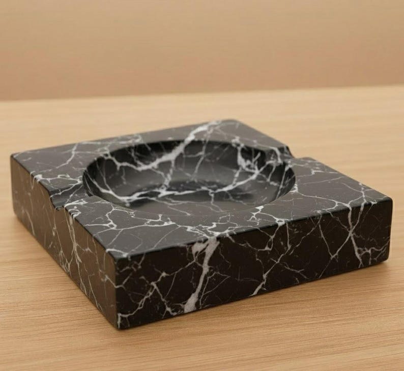 May include: A square ashtray made of black marble with white veining. The ashtray has a circular indentation for holding items and a rectangular shape. The ashtray is on a wooden surface.