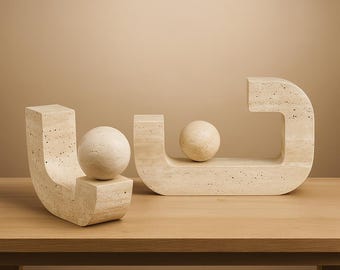 Puede incluir: Dos esculturas abstractas de piedra beige claro. Una tiene forma de U con una esfera, la otra una forma curva con una esfera. Las esculturas están sobre una superficie de madera.