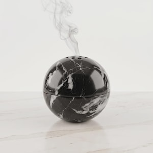 May include: A spherical black and white marble incense burner with a smooth, polished finish. The top half features a series of small circular vents, and a thin trail of grey smoke rises vertically from the center of the sphere's top surface.