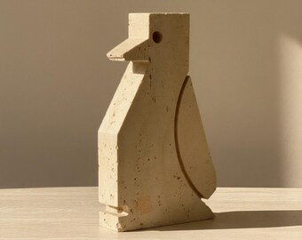 Könnte beinhalten: Eine beige, geometrische Vogelskulptur mit quadratischem Kopf, dreieckigem Schnabel und kreisförmiger Vertiefung an der Seite. Die Skulptur besteht aus einem strukturierten Material und steht auf einer hellen Oberfläche.