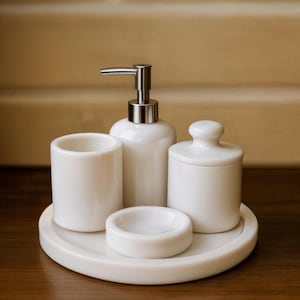 Può includere: Un set da bagno in ceramica bianca composto da un dispenser per sapone con pompa argentata, un bicchiere, un piattino e un barattolo con coperchio. Il set è presentato su un vassoio rotondo, il tutto in una finitura bianca lucida coordinata.