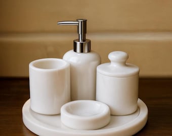 May include: A white ceramic bathroom set includes a soap dispenser with a silver pump, a tumbler, a small dish, and a jar with a lid. The set sits on a round tray, all in a matching glossy white finish. The background is a warm, neutral color.