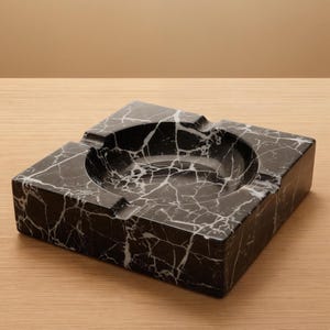 May include: A square ashtray made of black marble with white veining. The ashtray has a circular indentation in the center and a rectangular shape with a smooth, polished finish. The ashtray has a classic design.
