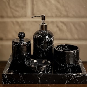 May include: A black marble bathroom set with white veining. The set includes a soap dispenser, a small jar with a lid, a tumbler, a small dish, and a rectangular tray. The dispenser has a silver pump.