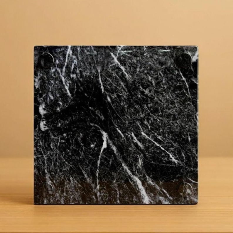May include: Square black marble coaster with white veining. The coaster has a polished finish and small, round feet on the bottom. The marble has a natural, textured appearance, and the coaster is set against a neutral background.