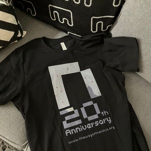 May include: A black short-sleeved t-shirt featuring a graphic print of a pixelated game level design. Below the graphic, the text reads 20th Anniversary in a blocky font, followed by the website address www.thewayoftheninja.org in white text.