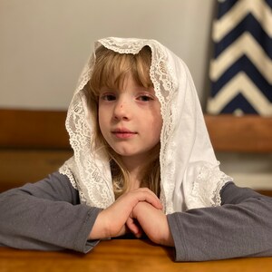 May include: A young person wearing a white lace head covering with a gray long-sleeved shirt. The person is sitting at a wooden table with their hands clasped together.