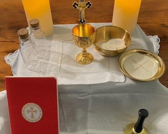 May include: A white tablecloth with a lace border, a red book with a gold cross, two clear glass bottles, two white candles, a gold chalice, a gold bowl with white wafers, a gold plate with a white wafer, and a gold bell with a black handle.