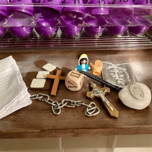 May include: A collection of religious items including a small wooden cross, a chain, a white stone with a religious figure, a small wooden figure of a woman in a blue dress, a wooden die with Roman numerals, a small wooden cross with a figure of Jesus, and a small white cloth with a religious design.