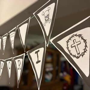 Op de afbeelding: Een zwart-wit banner met driehoekige vlaggen aan elkaar geregen. Sommige vlaggen tonen een schedel en gekruiste beenderen, een kruis in een krans en individuele letters die een woord spellen. De banner hangt tegen een neutrale achtergrond.