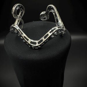 May include: A silver-toned bracelet with an openwork design. The bracelet has a V-shape with curved ends and a textured surface. The bracelet is displayed on a black surface against a dark background.