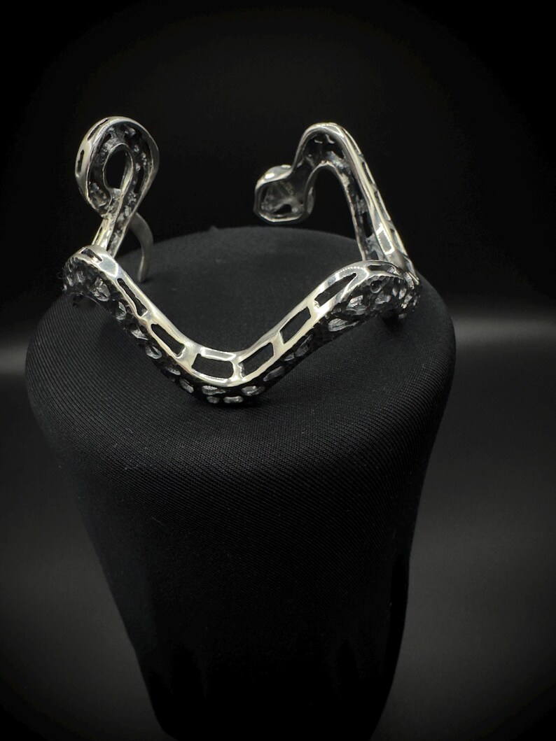 May include: A silver-toned bracelet with an openwork design. The bracelet has a V-shape with curved ends and a textured surface. The bracelet is displayed on a black surface against a dark background.