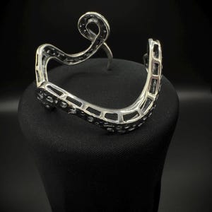 May include: A silver-toned cuff bracelet with an open design. The bracelet features a wavy, openwork pattern with a decorative swirl at the top. The cuff is displayed on a black surface against a dark background.