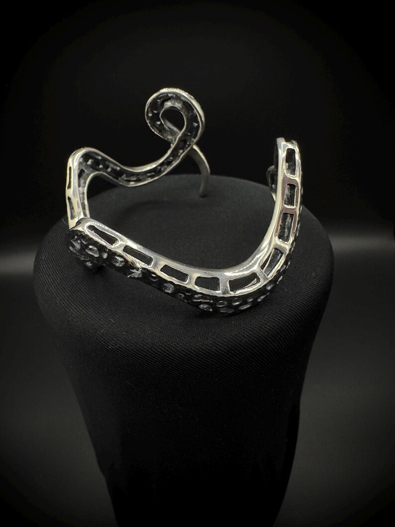 May include: A silver-toned cuff bracelet with an open design. The bracelet features a wavy, openwork pattern with a decorative swirl at the top. The cuff is displayed on a black surface against a dark background.