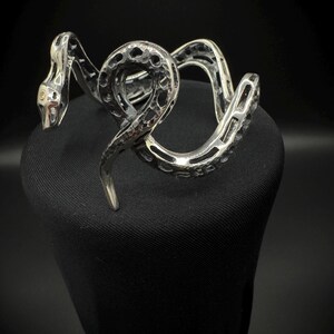 May include: A silver-toned snake bracelet with a textured, openwork design. The snake's head features a detailed eye, and the body coils around itself. The bracelet is displayed on a black surface, creating a contrast that highlights the intricate metalwork.