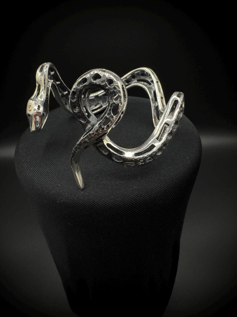 May include: A silver-toned snake bracelet with a textured, openwork design. The snake's head features a detailed eye, and the body coils around itself. The bracelet is displayed on a black surface, creating a contrast that highlights the intricate metalwork.