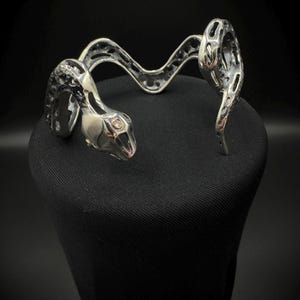 May include: A silver-toned snake-shaped cuff bracelet. The bracelet features a detailed snake head with an open mouth and a textured body. The design includes openwork patterns and a curved, undulating form, creating a striking accessory.