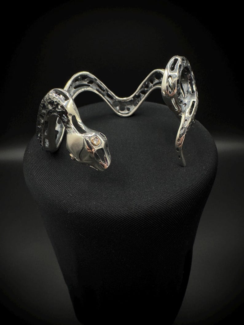 May include: A silver-toned snake-shaped cuff bracelet. The bracelet features a detailed snake head with an open mouth and a textured body. The design includes openwork patterns and a curved, undulating form, creating a striking accessory.