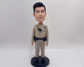 May include: A bobblehead figurine of a person wearing a tan uniform with a black belt and a black radio. The person has short black hair and is wearing a badge on their chest.