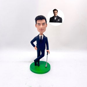 May include: A custom bobblehead figurine of a person wearing a dark blue suit and a white shirt with a tie. The figurine is holding a golf club and standing on a green base. The figurine is looking to the side and has a serious expression on their face.