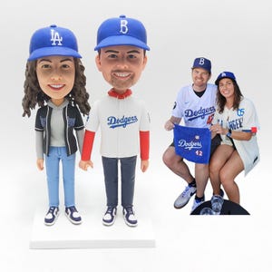 May include: Bobblehead figurines of a couple wearing blue baseball caps and Dodgers jerseys. The figurines are standing on a white base. A couple is sitting in the background, also wearing Dodgers apparel and holding a blue towel.