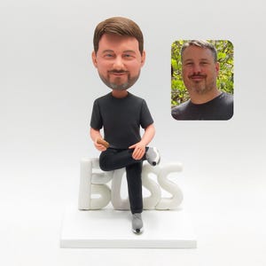 May include: A personalized bobblehead figurine of a man in a black t-shirt and pants, seated on a white base that spells out "BOSS". The figurine has a realistic head and a small photo of the person. The shoes are gray.