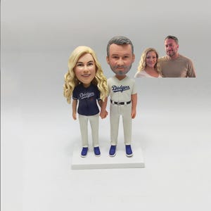 May include: A custom-made cake topper featuring a couple dressed in baseball uniforms. The woman is wearing a blue jersey with the word "Dodgers" on it, and the man is wearing a white jersey with the same word. They are both holding hands and smiling.