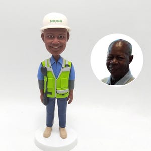 May include: A custom bobblehead figurine of a construction worker wearing a white hard hat with "MOSS" in green, a neon green safety vest, blue shirt, and jeans. A small photo of a man is in the background.