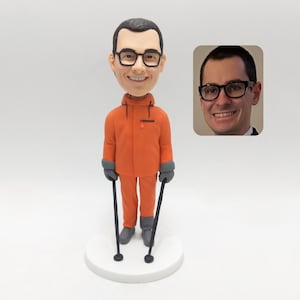 May include: A personalized bobblehead figurine of a man wearing an orange winter jacket, gray gloves, and gray boots, holding two black walking sticks. The figurine has a smiling face and glasses. A photo of the man is in the background.
