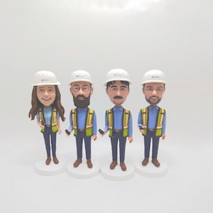May include: Four custom bobblehead figurines depicting construction workers wearing white hard hats, light blue button-down shirts, high-visibility yellow and orange safety vests, and dark blue trousers, each standing on a circular white base.