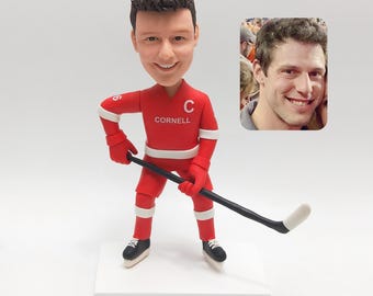 May include: A red hockey player figurine with a smiling face, wearing a red uniform with the word "Cornell" and the letter "C". The figurine is holding a black hockey stick with a white blade. The figurine is on a white base.