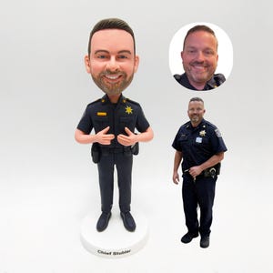 May include: A custom bobblehead figurine of a police officer in a dark blue uniform, with a smiling face and a badge. The figurine is standing on a white base with the name "Chief Stubler" printed on it. A photo of the person is in the background.