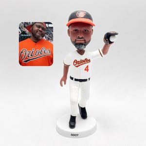 May include: A bobblehead figurine of a baseball player in a white uniform with the word "Orioles" and the number "4" on the front. The figurine is holding a baseball and wearing a black cap with an orange brim. A photo of a person wearing an Orioles jersey is in the background.