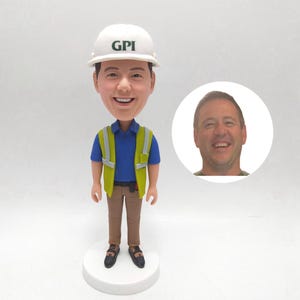 以下が含まれることがあります： 「GPI」とプリントされた白いヘルメットをかぶった、笑顔の男性の首振り人形。青いポロシャツ、黄色の安全ベスト、茶色のズボンを着用しています。人形は白い台座の上にあり、男性の顔の小さな画像が付いています。