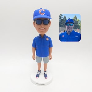 May include: A bobblehead figurine of a man wearing a blue baseball cap with a red "C" logo, sunglasses, a blue short-sleeved shirt, gray shorts, and sneakers. The figurine is on a white base with the text "Coach Adam". An inset photo shows the man.