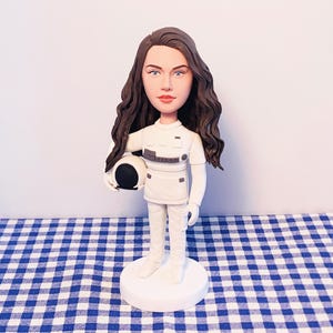May include: A figurine of a woman astronaut in a white uniform, holding a black and white helmet. The figure has long brown hair and is standing on a white circular base. The background is a blue and white checkered pattern.