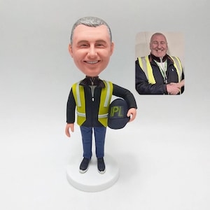 May include: A custom bobblehead figurine of a man wearing a black jacket, blue pants, and a yellow safety vest. The figurine is holding a navy blue hard hat with "PL" printed on it. A smaller photo of the man is in the background.
