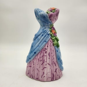 May include: A ceramic figurine in the shape of a dress. The dress features a light blue shawl with dark blue polka dots, a pink bodice, and a pink skirt with purple accents. The dress is adorned with floral and green leaf accents.