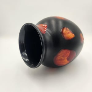 Puede incluir: Un jarrón de cerámica negro con un interior ancho y oscuro y un cuerpo redondeado. El jarrón presenta manchas irregulares de color rojo anaranjado en su superficie. El borde es liso y oscuro, contrastando con las manchas más claras. El jarrón está sobre un fondo blanco.
