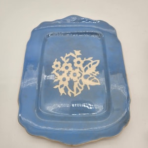 May include: A rectangular, blue ceramic serving tray with a scalloped edge. The tray features a white floral design in the center. The tray has a glossy finish and a light brown rim. A decorative piece for serving food or displaying items.