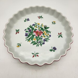 Puede incluir: Fuente de tarta de cerámica blanca con borde festoneado y diseño floral. El centro presenta un grupo de flores rosas, moradas, amarillas y verdes. Pequeños acentos florales están dispersos alrededor de la fuente, y el borde es rojo oscuro.