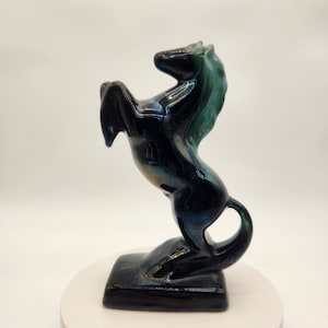 Op de afbeelding: Een keramieken paardenbeeldje in een steigerende pose. Het paard heeft een glanzende afwerking met een donkerblauwe body en een turquoise manen. Het beeldje is gemonteerd op een rechthoekige basis.