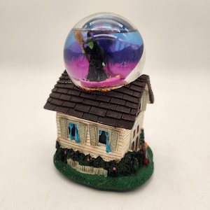 May include: A decorative snow globe featuring a green-skinned witch holding a broomstick, set atop a miniature house with a brown shingled roof, white siding, blue curtains, and a green base with dark green shrubbery and a small white picket fence.